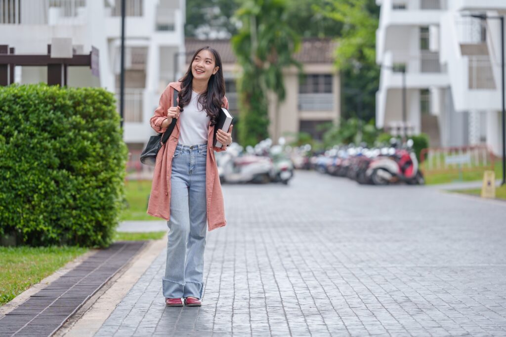 Cheerful asian student walks along a campus pathway, carrying books and a backpack, enjoying the ple
